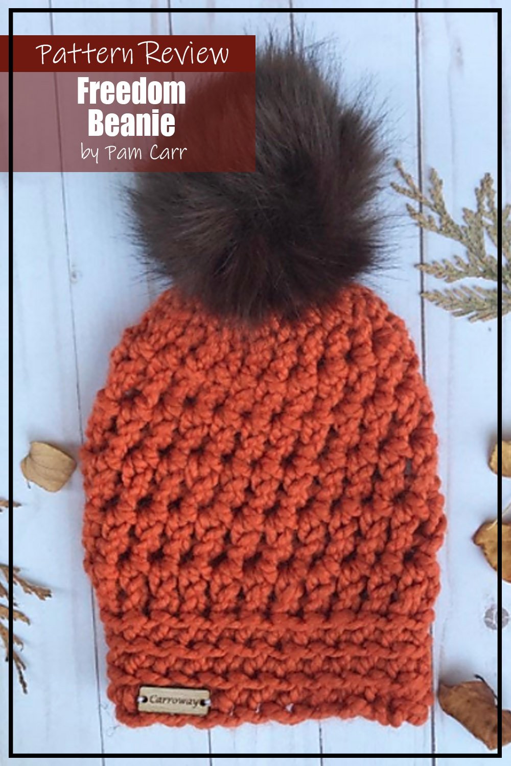 Freedom Beanie crochet hat pattern sample made with orange yarn and a brown furry pompom.  This crochet sample project is laying flat on a white wooden background.  Text on the image says "Freedom Beanie by Pam Carr Pattern Review".