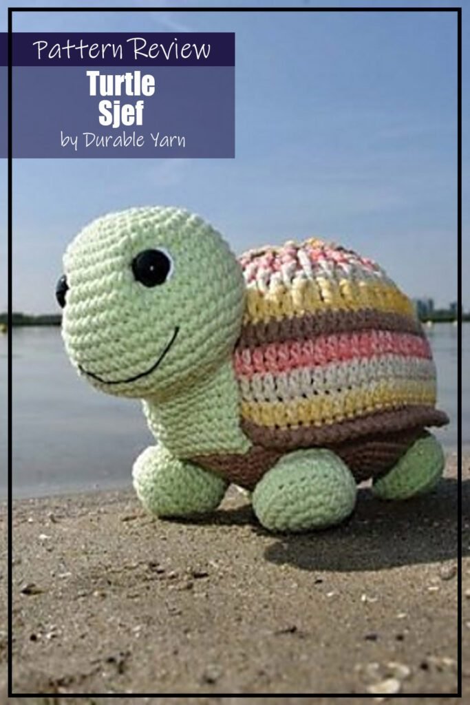 Turtle Sjef crochet pattern sample is a toy amigurumi turtle made with assorted colours on the shell, green head and feet, and a brown belly. The crochet project is sitting on a beach in front of water. The text on the image says "Turtle Sjef by Durable Yarn Pattern Review".