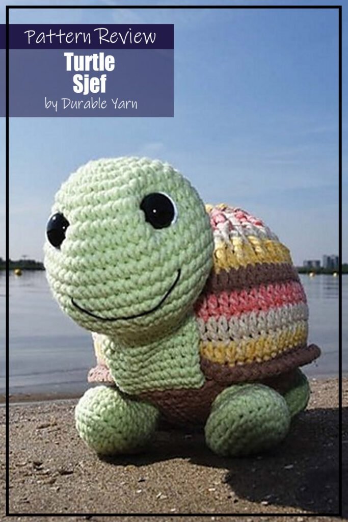 Turtle Sjef crochet pattern sample is a toy amigurumi turtle made with assorted colours on the shell, green head and feet, and a brown belly. The crochet project is sitting on a beach in front of water. The text on the image says "Turtle Sjef by Durable Yarn Pattern Review".