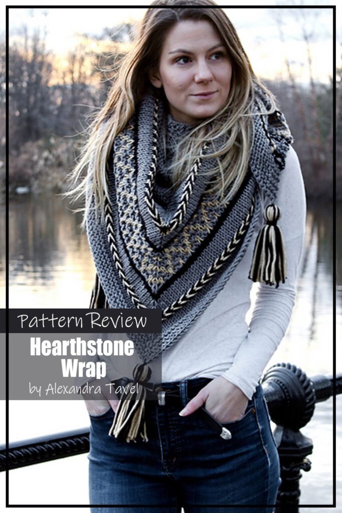 Hearthstone Wrap by Alexandra Tavel knitting pattern sample made with a dark grey yarn for the main body with accents of black and cream.
Modeled on a woman wearing jeans and a white long sleeve shirt, the picture shows the wrap tied around her neck as a scarf.
Text on the image says "Hearthstone Wrap by Alexandra Taval Pattern Review"