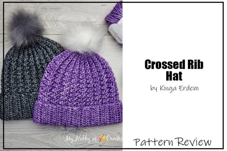 The Crossed Rib Hat crochet pattern samples are laying flat on a grey wood surface. One hat is a tonal dark grey aran weight yarn with a grey pompom and the other hat is a tonal purple yarn with a white pompom. The hat features a folded over ribbed brim with an interesting 3D ribbing on the main part of the hat. The text on the image reads "Crossed Rib Hat by Kinga Erdem Pattern Review".