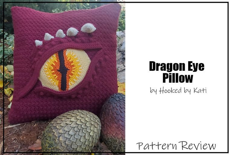 Dragon Eye Pillow crochet pattern sample is made with a burgundy yarn for the main pillow with accents of white, yellow, orange, and black for the dragon eye. The pillow has scaley dragon eggs sitting in front of it on the lower right corner. The text on the image reads "Dragon Eye Pillow by Hooked by Kati Pattern Review".