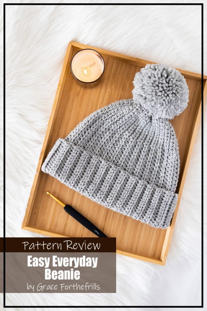 Easy Everyday Beanie crochet hat pattern sample is made using a grey wool. The hat features a ribbed texture and folded brim. The hat is laid flat on a wooden tray with a crochet hook before it and a candle above it. The text on the image reads "Easy Everyday Beanie by Grace Forthefrills Pattern Review".