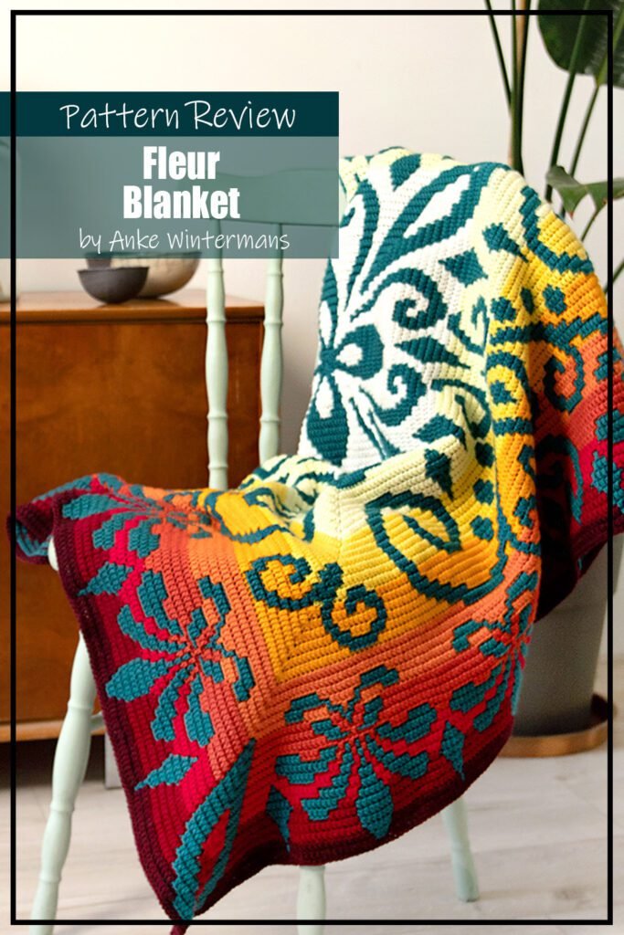 Fleur blanket crochet pattern sample is made using multiple yarn colours. The background is a gradiant of white through yellow, orange and then red. The decorative design is made with a rich green/teal coloured yarn and features a flower motif. The blanket is draped over a wooden chair. The text on the image reads "Fleur Blanket by Anke Wintermans Pattern Review".