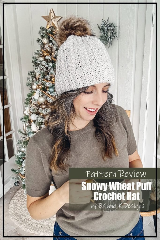 the Snowy Wheat Puff Crochet Hat pattern sample is made using a light grey worsted weight yarn. The beanie/toque has a ribbed brim and texture on the main part of the hat. The hat, which has a brown pompom on top, is modeled by a woman with brown hair. The text on the image reads "Snowy Wheat Puff Crochet Hat by Briana K Designs Pattern Review".
