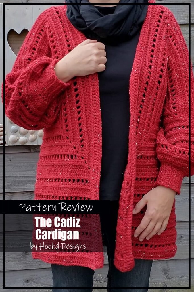 The Cadiz Cardigan crochet pattern sample is made using bright red yarn and modeled on a woman wearing black underneath which makes the texture of the cardigan stand out. The text on the image reads "The Cadiz Cardigan by Hookd Designs Pattern Review".