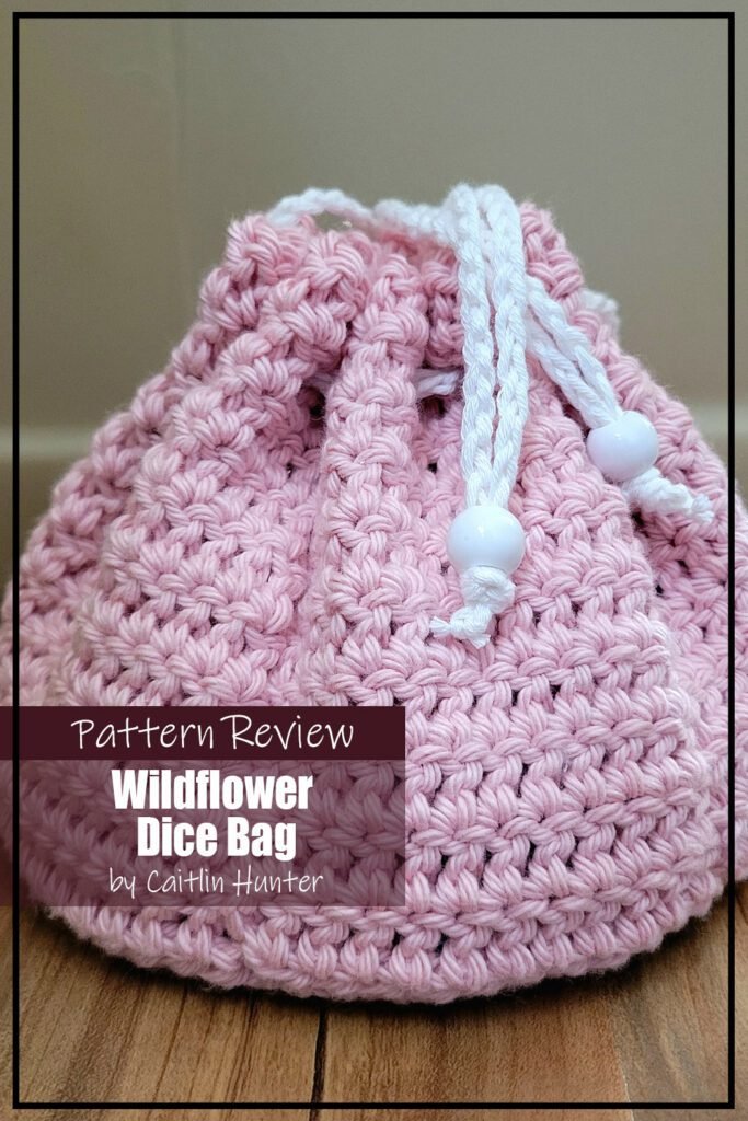 Wildflower Dice Bag crochet pattern sample is made with pink yarn an a white yarn tie. The bag is pulled closed and is sitting on a wooden surface. The text on the image reads "Wildflower Dice Bag by Alyssa P Pattern Review".