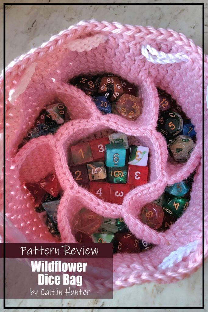 Wildflower Dice Bag crochet pattern sample is made with pink yarn an a white yarn tie. The bag is open showing the different compartments inside that are filled with an assortment of dice. The text on the image reads "Wildflower Dice Bag by Alyssa P Pattern Review".